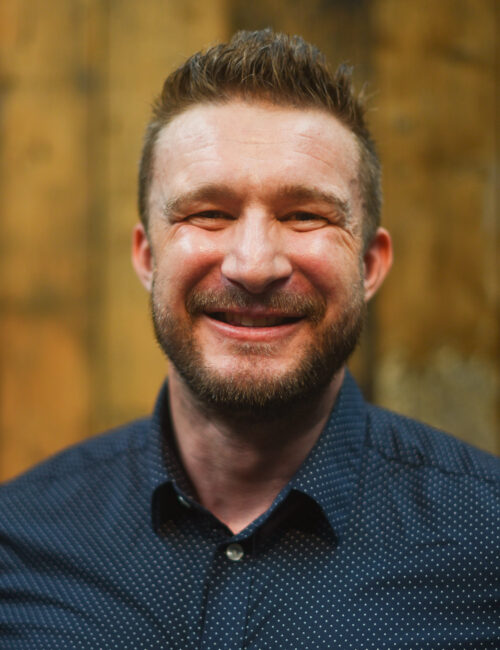 A headshot image of Alex Crawford smiling towards the camera.