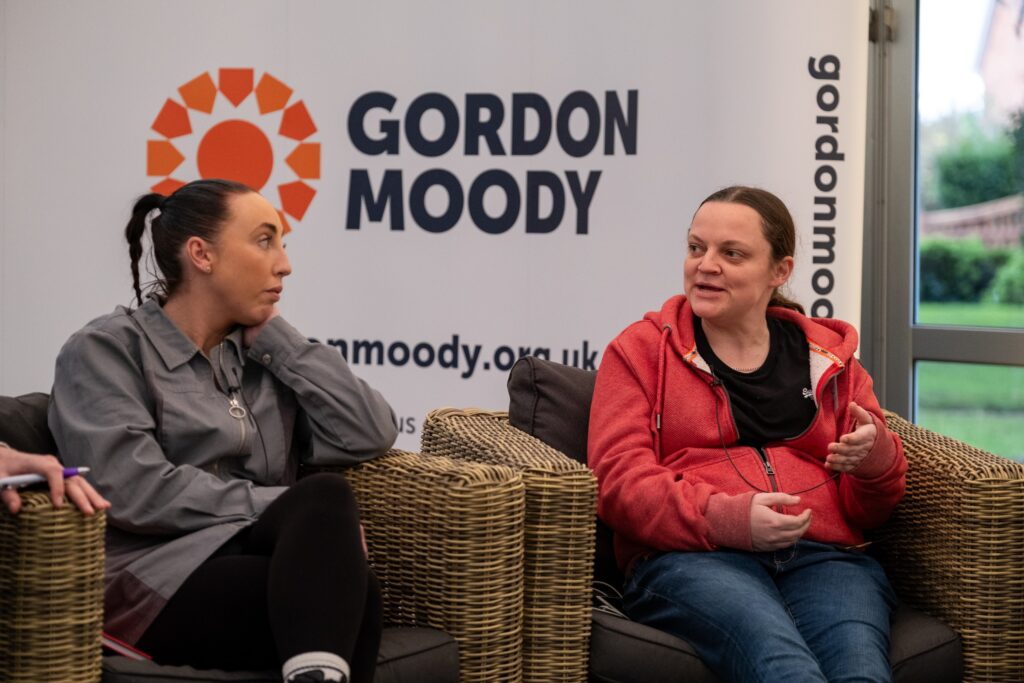 Two female Gordon Moody alumni, one wearing a grey blouse and one wearing an orange hoodie, speak on stage at the official opening event for Gordon Moody House in Redditch on Monday 25th November 2024.