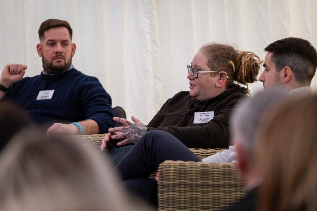 A female Gordon Moody alumni wearing a brown hoodie speaks on stage at the official opening event for Gordon Moody House in Redditch on Monday 25th November 2024. Also pictured are two male alumni, one in a blue jumper and one in a grey jumper.