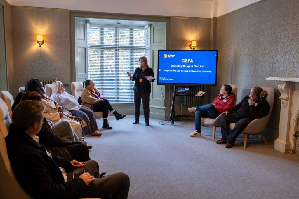 Gordon Moody Training Services Manager, Lisa Hartland, stands in front of a screen showing the title page of Gambling Support First Aid. Lisa is presenting to an audience of invited guests at the official opening of Gordon Moody House on Monday 25th November 2024.